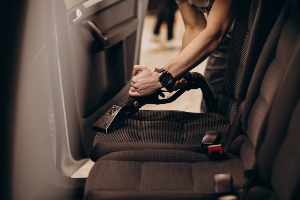 Person vacuuming fabric seats in a car interior using a specialized vacuum attachment.