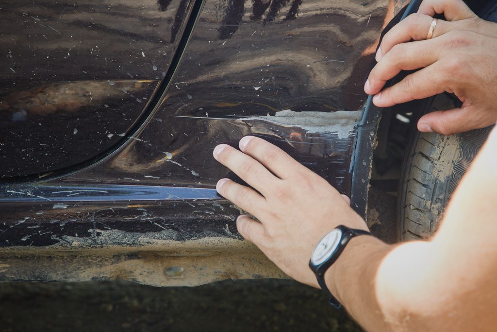 A person inspecting deep scratches and paint damage on the side panel of a black car with their hands.