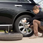 A driver learning how to change a car tire with a lug wrench and scissor jack.
